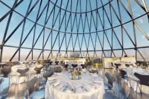 A modern, glass-domed dining room set for an event, with round tables, white tablecloths, and panoramic city views through large triangular windows. Consensus