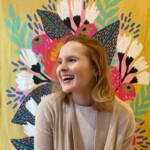 Sarah Frazier, with shoulder-length hair and a light sweater, smiles warmly in front of a vibrant floral mural adorned with pink, white, and green elements.