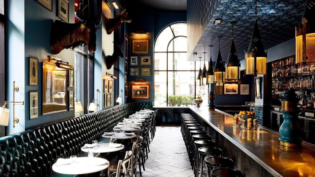 A dimly lit, stylish bar with high ceilings, bar stools, and tables along a cushioned bench. Walls are decorated with framed art and mounted animals. Consensus