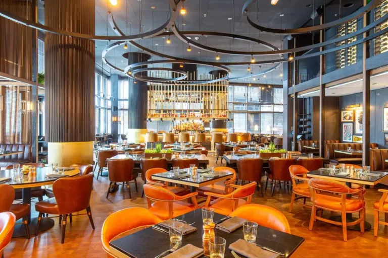 A modern restaurant with brown leather chairs, black tables set with glasses and napkins, large windows, and a central bar with bottles and hanging lights. Consensus