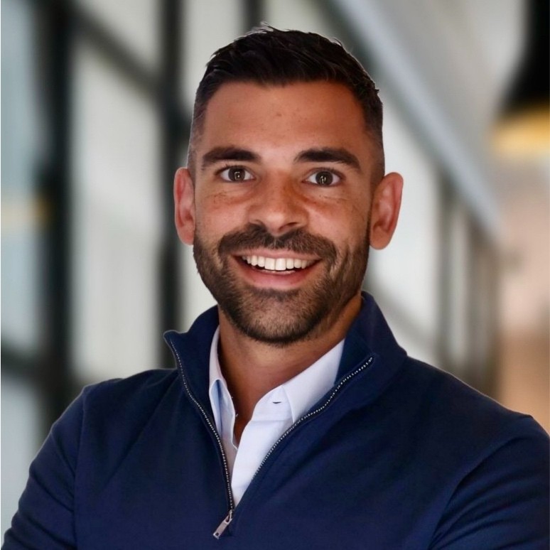 A person with short dark hair and a beard smiles, wearing a navy jacket over a white shirt. The background is a blurred indoor setting with large windows. Consensus