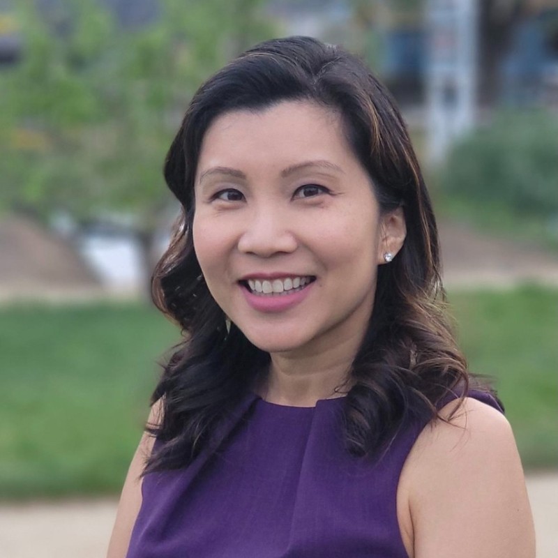 Betty Mok, with her long, wavy hair, smiles brightly in a purple dress. The background showcases a blur of lush greenery, enhancing the serene outdoor ambiance.