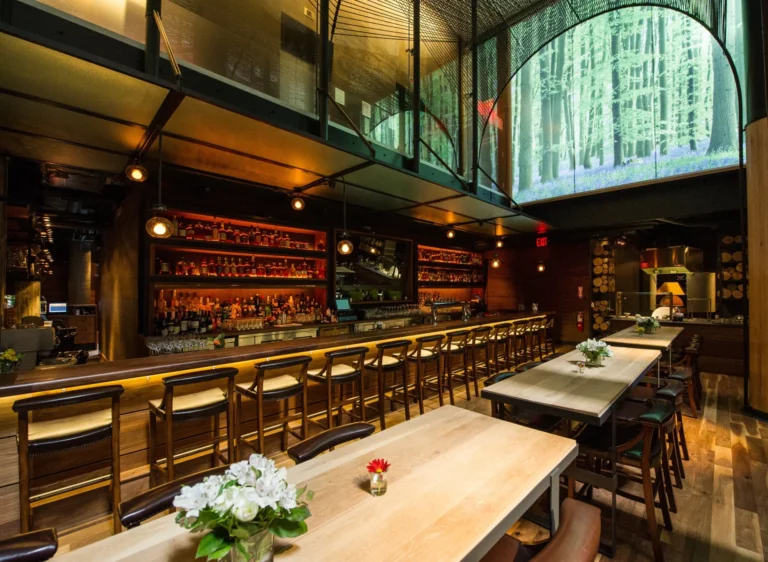 A dimly lit bar with a wooden counter, stools, and two long tables with flower arrangements. A large digital screen above displays a forest scene. Consensus