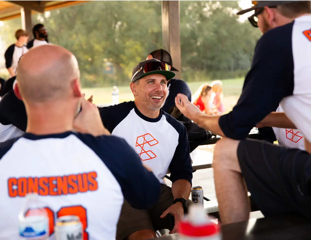 Man in a baseball cap smiling and conversing with teammates at a picnic table, all wearing jerseys with the word "consensus.