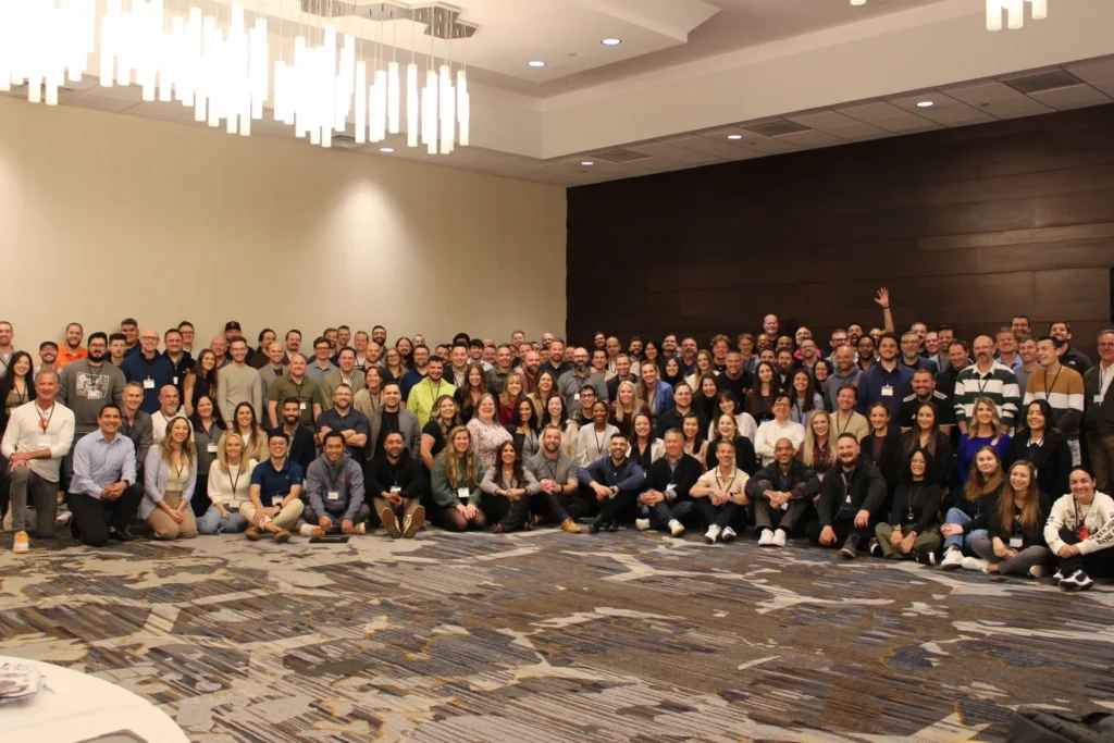 A large group of people posing together in a conference room with chandeliers overhead, smiling at the camera. Consensus