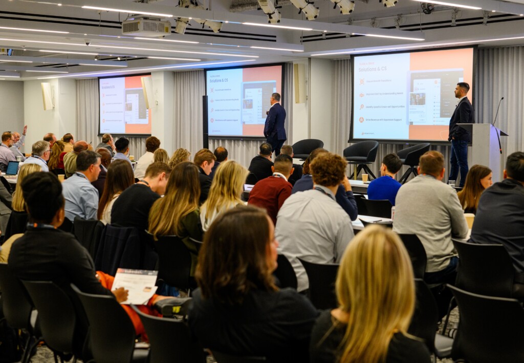 At DEMOFESTx on the East Coast, a speaker captivates a seated audience in the conference room, using two large screens to bring the slideshow presentation to life. Consensus