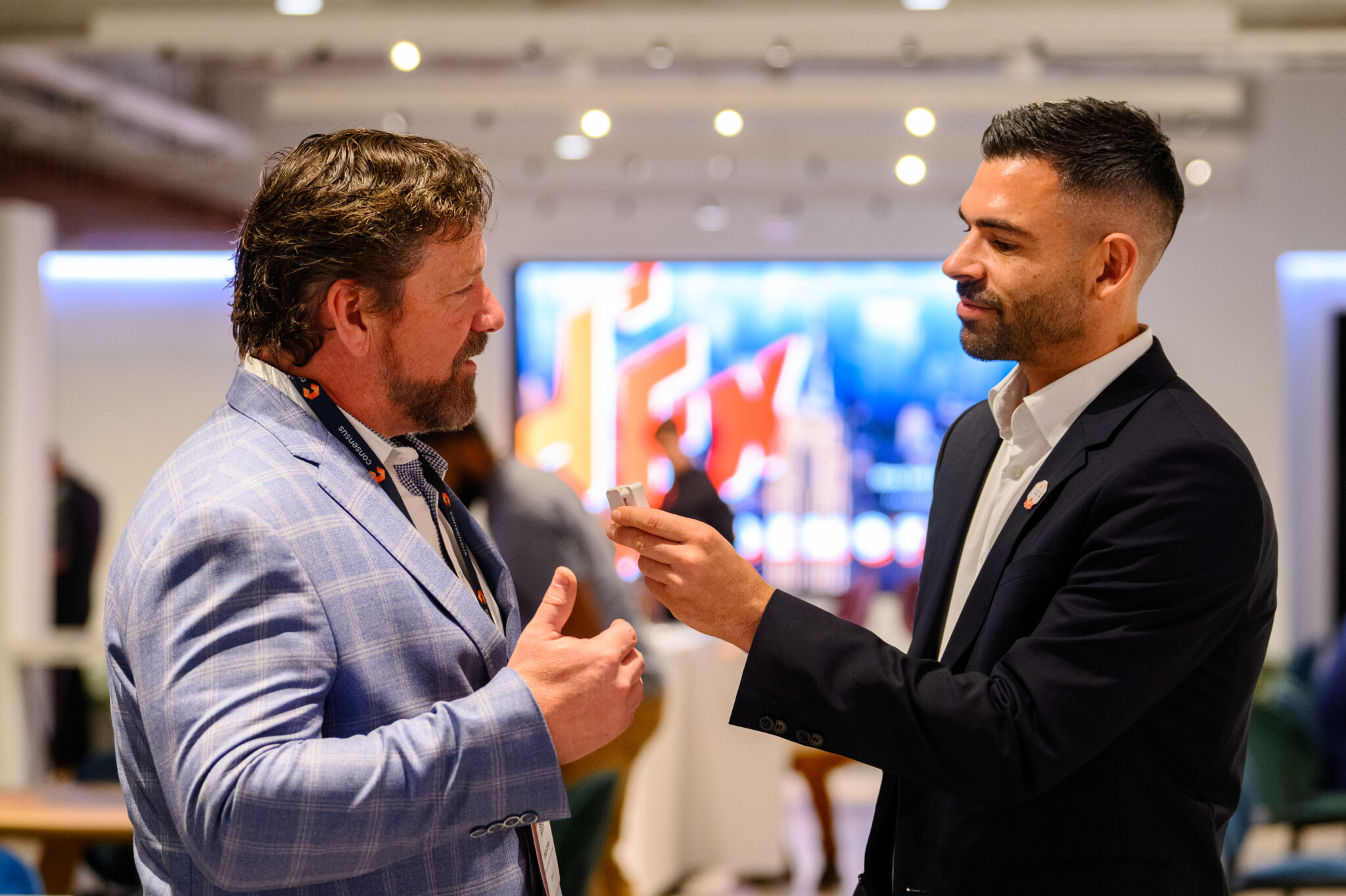 In a business setting on the East Coast, two men are deep in conversation. One grips a microphone, hinting they might be at DEMOFESTx. Behind them, a blur of colorful screens sets the scene for innovation and ideas. Consensus