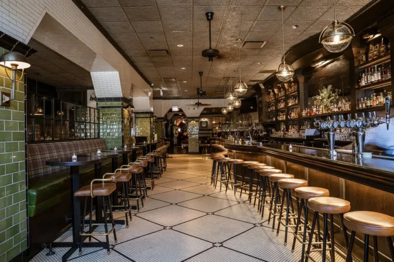Spacious bar interior with rows of wooden stools, a long counter, tiled floor, and shelves of liquor bottles behind the bar. The space appears empty and well-lit. Consensus