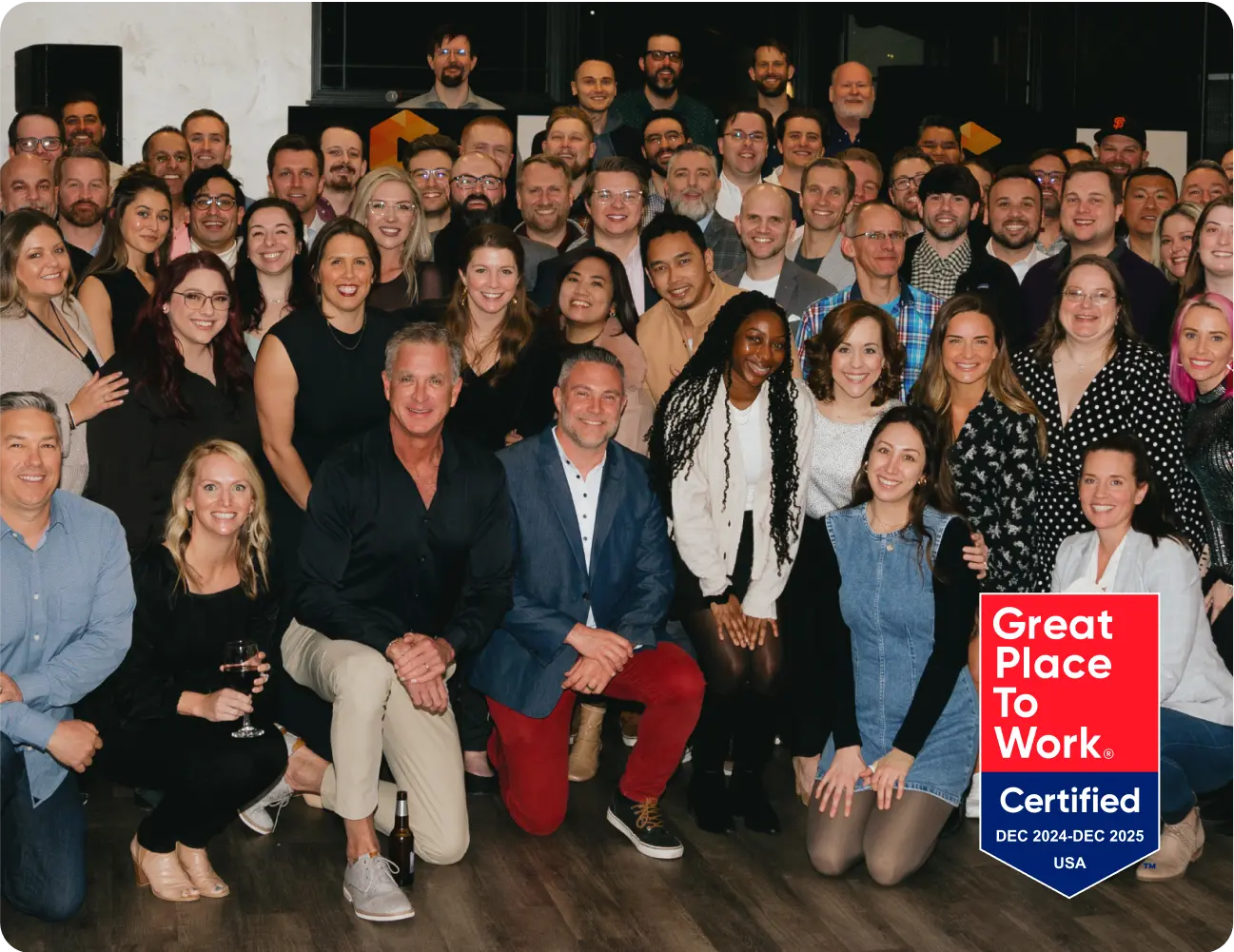 A large group of people posing together at an indoor event with a "Great Place to Work" certification badge in the corner. Consensus