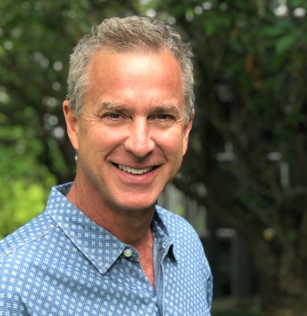 Doug Johnson, a middle-aged man with short gray hair, smiles in a blue patterned shirt while standing outdoors with green trees in the background. Consensus