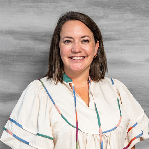 A woman in a white blouse with colorful ribbons smiles at the camera against a gray background.