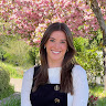 A woman with long brown hair smiles at the camera while standing outside in front of a tree with pink blossoms. She is wearing a white shirt under a black pinafore.