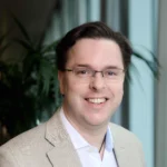 A man with short dark hair and glasses, identified as Mark Green, smiles at the camera while wearing a beige blazer over a light blue shirt. A blurred indoor background with a plant is visible behind him.