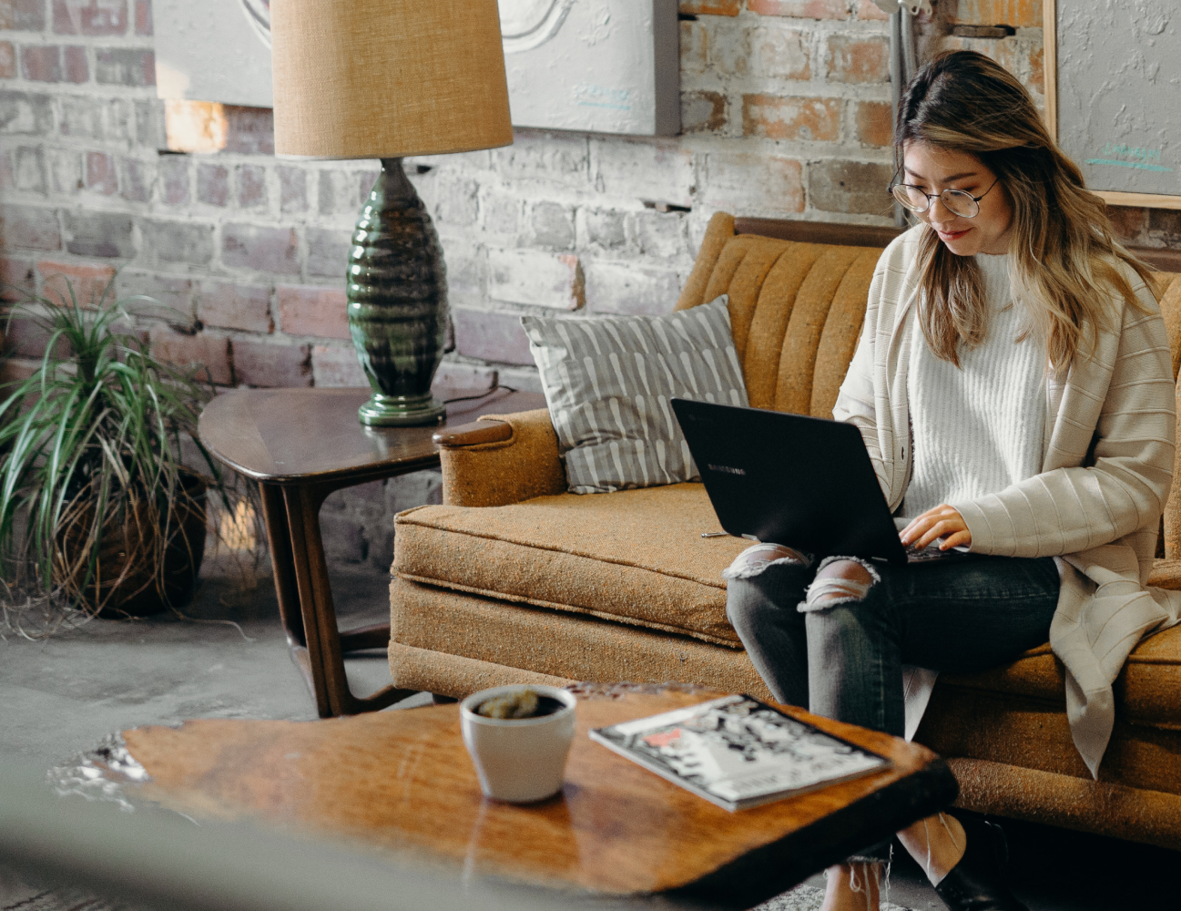 A woman sits on a brown sofa in a cozy living room, working on a laptop. A coffee table with a magazine and a potted plant are in the foreground. An exposed brick wall is in the background.
