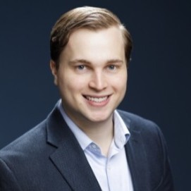 A man with short brown hair smiles, wearing a gray blazer over a light blue shirt in front of a dark background, showcasing his confident yet approachable demeanor.