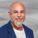 Bald man with a gray beard wearing a blue blazer and white shirt, smiling in front of a gray background—this is Mike Trionfo. Consensus