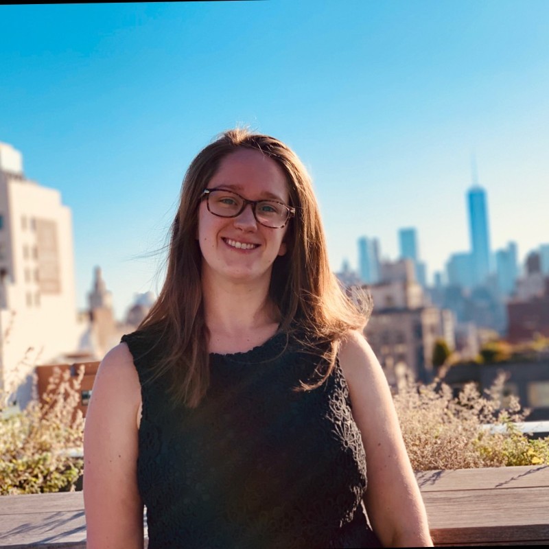 A woman with glasses and long hair smiles while standing on a rooftop, the city skyline stretching out behind her on a clear day. Her confident gaze suggests expertise in sales efficiency.