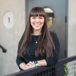 Melissa Megginson, with her long brown hair and black top, smiles warmly while standing in front of a round window. Her outfit is complemented by a striking green watch that catches the eye. Consensus