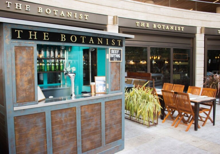 Outdoor bar named "the botanist" with empty wooden chairs and a plant nearby, featuring beer taps and open shelving.