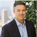 A man in a dark suit and checkered shirt stands in front of a leafy plant with a cityscape and tower visible in the background. Consensus