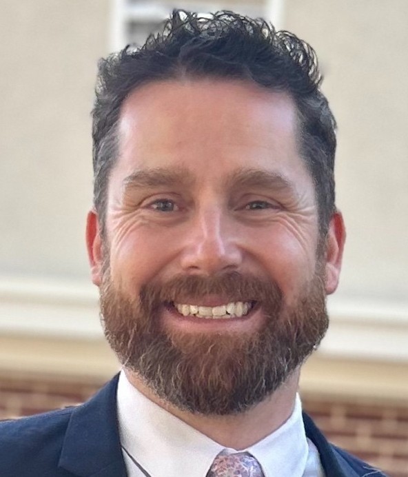 A man with curly dark hair and a full beard is smiling, wearing a suit and tie, standing outdoors in front of a blurred building background. Consensus
