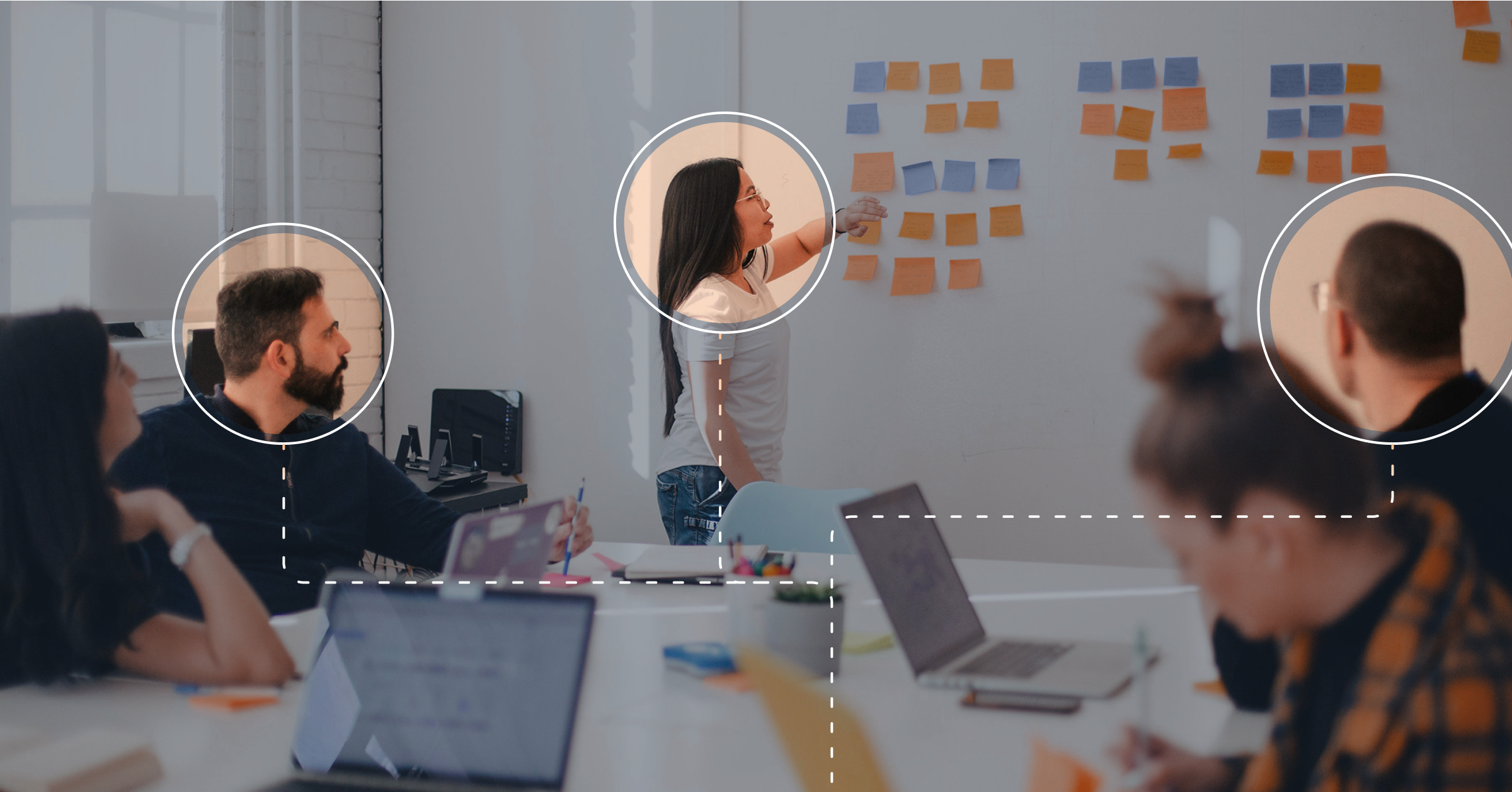 Four people work in an office. One person is placing sticky notes on a whiteboard, identifying and targeting key sales stakeholders. Two people are seated, facing each other, while another person works on a laptop.