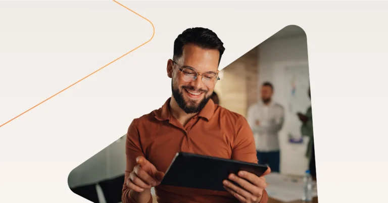 A man with glasses and a beard, wearing a rust-colored shirt, smiles while using a tablet—capturing the essence of Sales Enablement. Two people stand in the blurred background of an office setting. Consensus