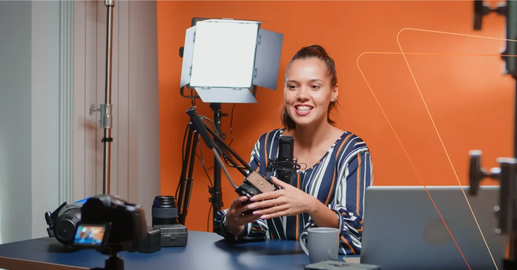A woman in a recording studio talks into a microphone. She is holding a tablet with various recording equipment around her, capturing every detail of the customer journey. An orange wall is in the background.