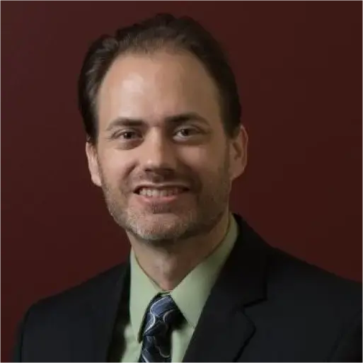 A man in a dark suit, green shirt, and patterned tie—reminiscent of Ken Connally's signature style—smiles against a dark red background.