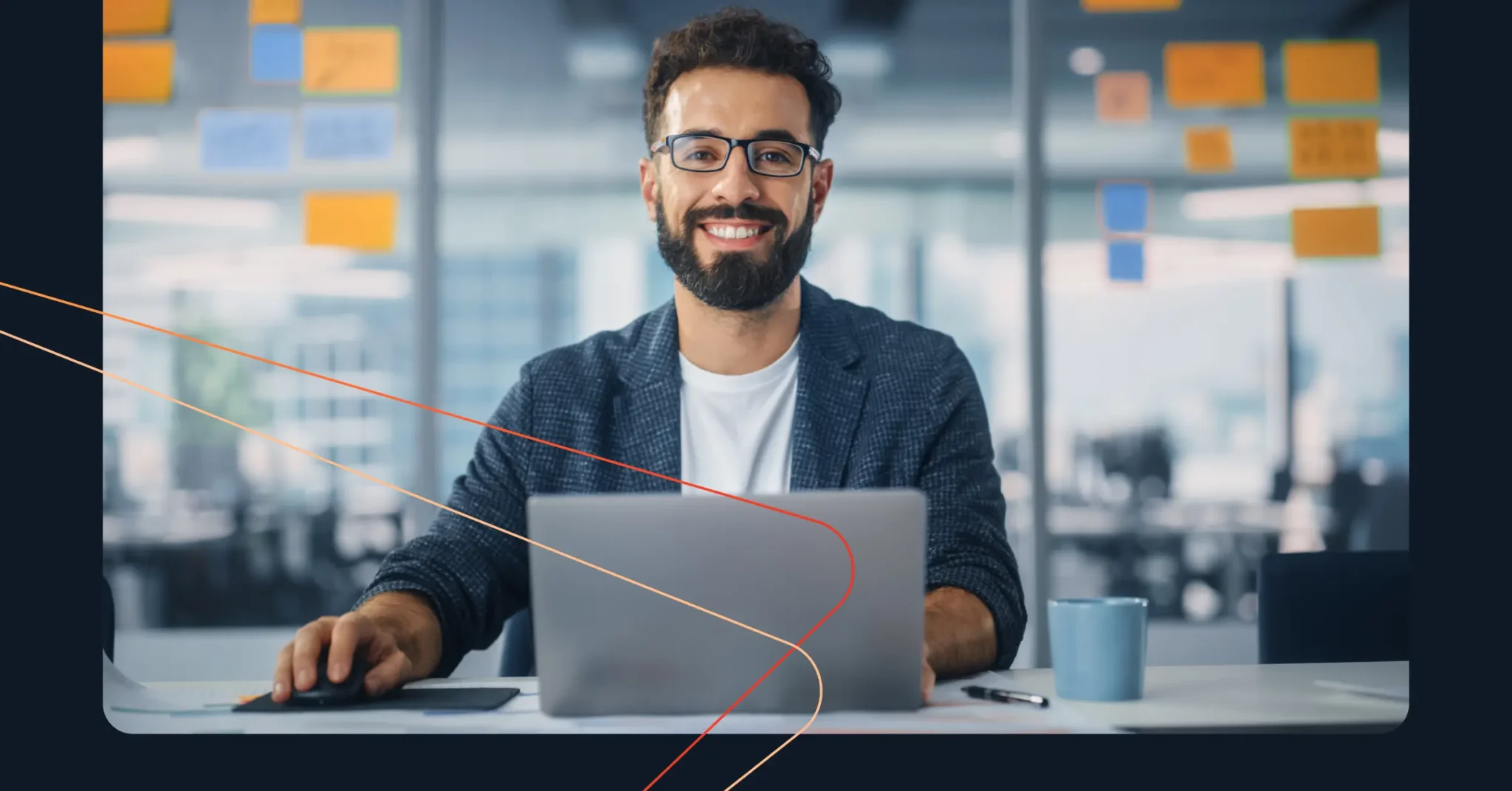 A man with glasses and a beard smiles while working on a laptop at a desk, refining his sales strategy. He holds a computer mouse, with a blue mug nearby. Office setting with sticky notes on the glass wall in the background.