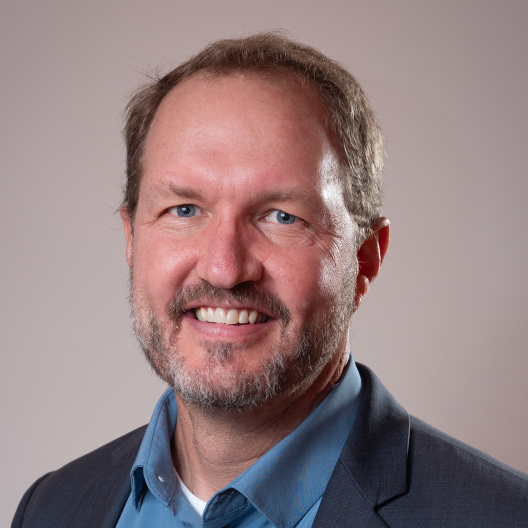 A professional headshot of a smiling middle-aged man with a beard, wearing a blue shirt and dark blazer, against a neutral background.