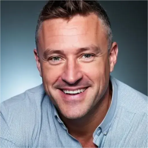 Tod Goulds, a man with short hair, wearing a light blue button-up shirt, smiles warmly at the camera against a blue background.