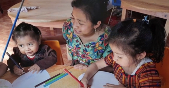 Three children at a wooden table engaged in charity activities; the youngest looks at the camera, the other two focus on coloring with pencils.