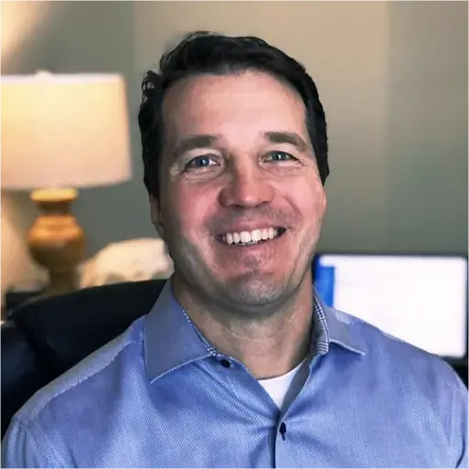 John Hirsch, in a blue shirt, smiles warmly indoors, framed by a softly glowing lamp and the subtle light of a computer screen in the background.