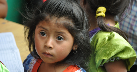 Young girl with a thoughtful expression, wearing a green blouse and her hair tied in a ponytail, surrounded by other children in a charity classroom setting.