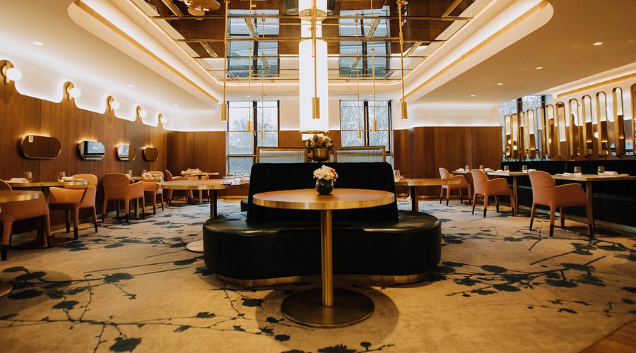 A modern, upscale restaurant interior with round tables, upholstered chairs, large windows, and contemporary lighting. The space features wood accents and floral-patterned carpet. Consensus