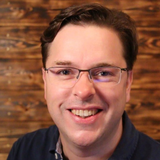 A smiling man with glasses, wearing a blue shirt, posing in front of a wooden panel background.