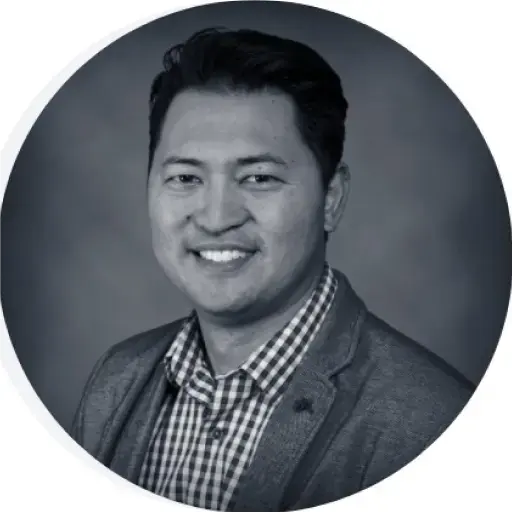 Portrait of a smiling person, possibly Keith Tanaka, in a checked shirt and blazer, looking at the camera against a gray background.